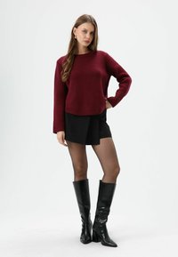Bordeauxrode gebreide trui met lange mouwen, gecombineerd met een zwarte geplooide minirok en zwarte knielange laarzen met hak.