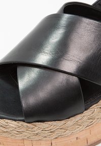 Černá kožená sandál s širokými křížícími se pásky, espadrilová podrážka z pletené jutové příze s hladkým povrchem a zaobleným tvarem.