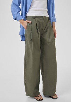 Személy olívazöld bőszárú nadrágot, fehér ujjatlan felsőt, kék feltűrt inget és fekete strandpapucsot visel, egyik kezével a zsebében.