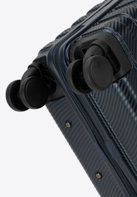 Eckbereich eines blauen Carbonfaser-Koffers mit zwei schwarzen Rädern, passenden Reißverschlüssen und silbernem Zubehör, das eine strukturierte Oberfläche und einen glatten Finish aufweist.