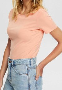 Pfirsichfarbene, kurzärmlige T-Shirts aus glattem Material, kombiniert mit hellblauen, hoch taillierten Jeans-Shorts mit Knopfverschluss.