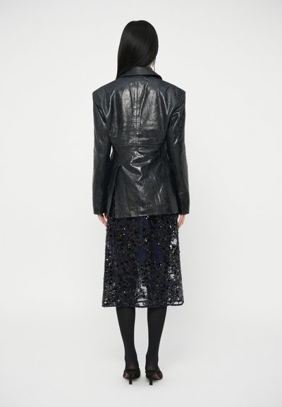 Blazer preto em couro com ombros estruturados, combinado com uma saia de lantejoulas azul-marinho até ao joelho. Modelo usa collants pretos e saltos altos.