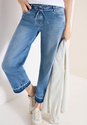 Kvinde iført lyseblå jeans i cropped model med stofbælte, hvide og blå sneakers, holder en hvid skjorte med smalle striber.
