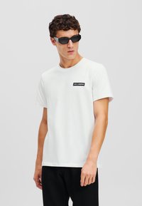Tricou alb din bumbac, cu guler rotund și mâneci scurte. Are o etichetă neagră cu text alb pe piept. Este purtat cu pantaloni negri și ochelari de soare.