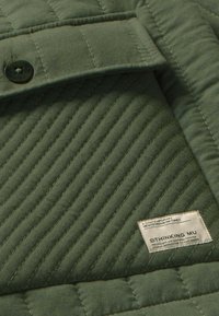 Gequiltede groene stof met een textuurpatroon en een zwarte knop. Inclusief een label met "THINKING MU" en productdetails in het wit.