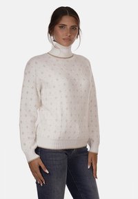 Maglione bianco a maglia con un collo alto a coste, caratterizzato da un motivo testurizzato e sottili accenti beige. Polsini e orlo a coste. Indossato con jeans.