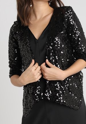 Femme portant une veste noire à sequins sur une robe noire, ajustant la veste avec les deux mains près de la poitrine.