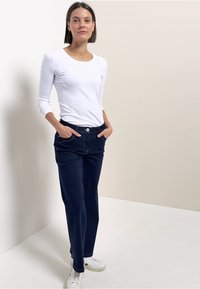 Parte superior blanca de manga larga combinada con pantalones azul marino. La modelo está de pie con las manos en los bolsillos. El calzado son zapatillas blancas. Líneas simples y limpias.