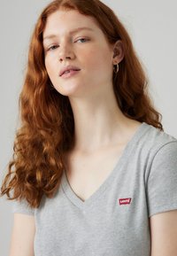 Gray short-sleeve V-neck t-shirt made of soft fabric, featuring a red logo label on the left side. Model has curly red hair.