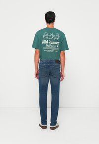 Camiseta de algodón verde con texto gráfico en blanco e ilustraciones en la parte posterior, combinada con jeans de mezclilla azules y zapatos marrones.
