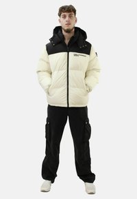 Giacca bianca gonfia con dettagli neri, cappuccio e zip frontale. Pantaloni cargo neri con tasche laterali, abbinati a sneakers bianche.