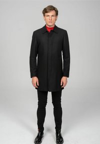 Zwarte wollen jas met een kraag, een sluiting aan de voorkant en lange mouwen, gedragen over een rode coltrui, gecombineerd met zwarte slim-fit broek en laarzen.