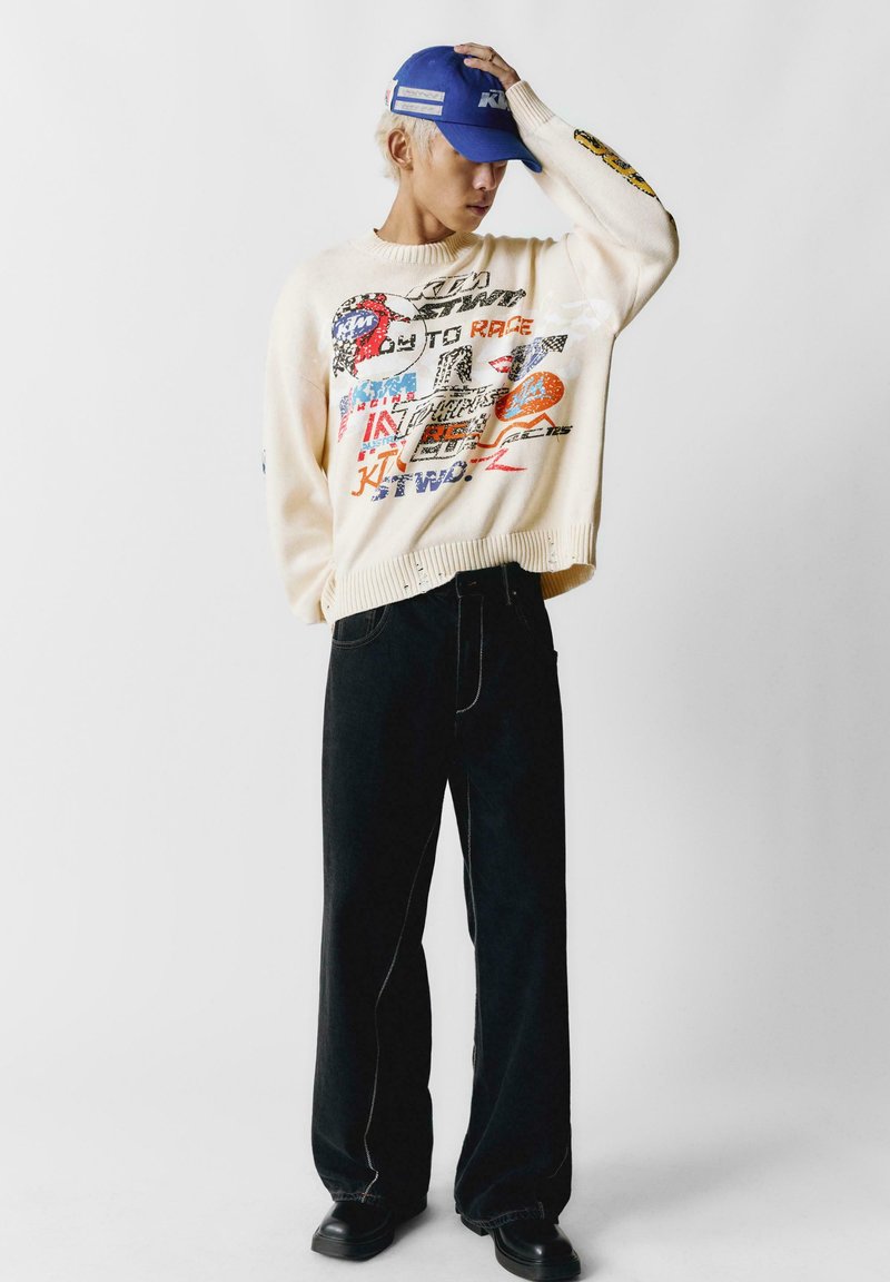 Persona che indossa un maglione grafico color crema, pantaloni neri a gamba larga, scarpe nere e un cappellino blu, posando con una mano sul cappellino.