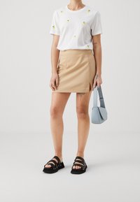 Bílý tričko s žlutou výšivkou citronu, béžová sukně s bočním rozparkem, černé sandály a malá modrá kabelka. Hladké textury po celém outfitu.