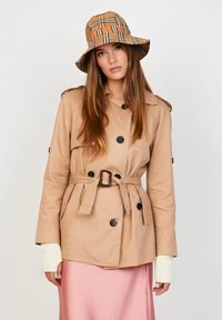 Abrigo trench beige con botones de doble fila, un cinturón en la cintura y hombreras en los hombros, combinado con un sombrero de cubo de cuadros marrón.