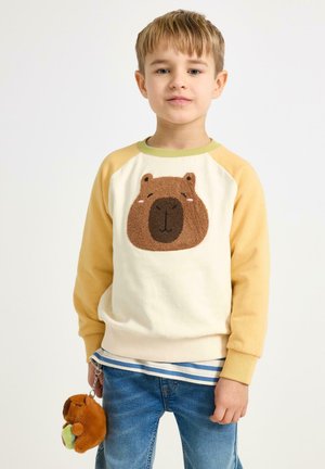 Crèmewitte sweatshirt met gele raglanmouwen en een bruine berenkop als applicatie. Blauwe spijkerbroek met een gestreept shirt dat eronder zichtbaar is. Een klein knuffeltje hangt aan de zak.