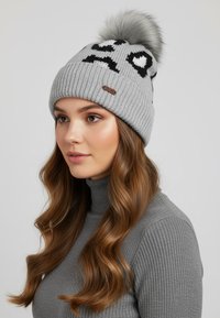 Gray knitted beanie with a fluffy pom-pom on top, featuring black designs including a heart and facial expressions, and a leather tag accent.