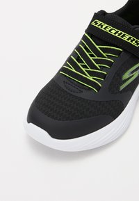 Zapatilla deportiva negra con parte superior de malla texturizada y cordones verde brillante. Cuenta con una suela blanca y una correa negra con la marca "SKECHERS".