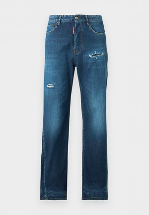 Άνδρας που στέκεται με το ένα χέρι στην τσέπη, φοράει λευκό φανελάκι, μπλε τζιν με ελαφρές φθορές και μαύρα παπούτσια σε απλό φωτεινό φόντο.