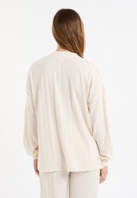 Cremefarbenes, geripptes Strickoberteil mit langen Ärmeln, lockerem Schnitt und rundem Ausschnitt, von hinten gezeigt, mit strukturierten Details.