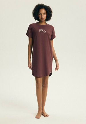 Kobieta o kręconych włosach, stojąca boso, ubrana w krótką sukienkę z krótkim rękawem do kolan, w kolorze brązowym, z drobnymi białymi grafikami owoców na piersi.