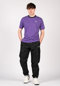 Violet t-krekls ar melnu apkakli un sānu apdari, apvienots ar melnām biksēm ar zaļu logo, valkāts ar baltām kedām.