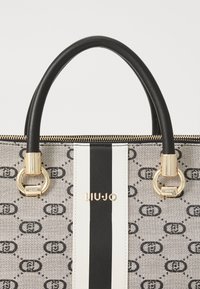 Beige designer tote bag with a patterned exterior, black leather handles, white and black striped accent, and gold hardware detailing.