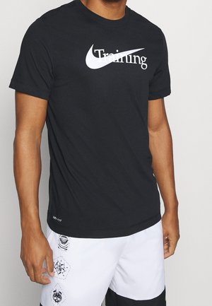 Hombre vistiendo una camiseta negra Nike Dri-FIT con el texto "Entrenamiento" y el logo de swoosh en blanco, combinada con pantalones cortos blancos que presentan parches gráficos en negro.