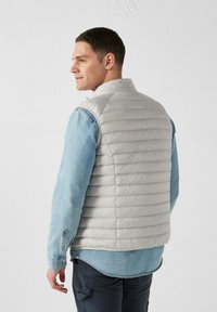 Gilet imbottito grigio con strisce verticali, indossato sopra una camicia in denim azzurro chiaro. Presenta un colletto alto e una consistenza liscia.
