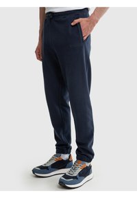 Vyras vilki tamsiai mėlynas sportines kelnes su elastinėmis apačiomis ir raišteliais, ranka kišenėje, kartu dėvi mėlynos ir rudos spalvų sportinius batelius bei baltas kojines.