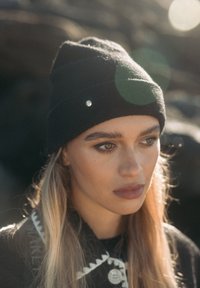 Zwart beanie-muts met een zilveren accent, aansluitend ontwerp, gemaakt van zacht materiaal. Op de achtergrond is een vervaagde rotsachtige textuur zichtbaar.