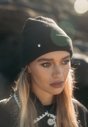 Chapeau en laine noire avec un accent argenté, design ajusté, fabriqué en matériau doux. En arrière-plan, une texture rocheuse floue est visible.
