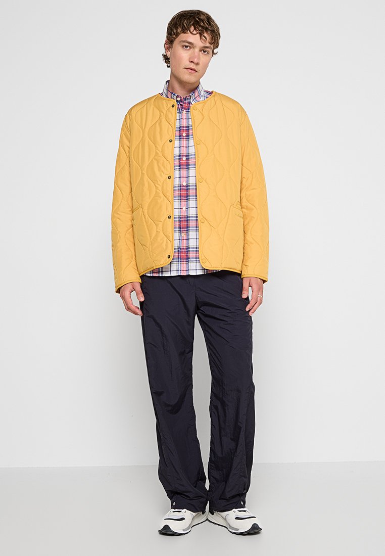 Gelbe gesteppte Jacke mit abgerundetem Saum und Druckknopfverschlüssen, kombiniert mit einem blau-roten karierten Hemd, dunkelblauen Hosen und weißen Sneakern.
