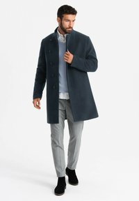 Cappotto di lana blu navy con taglio dritto, caratterizzato da un colletto alto e chiusura con cinque bottoni. Abbinato a un maglione azzurro chiaro, pantaloni grigi e stivali neri.