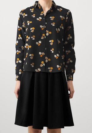 Czarna bluzka zapinana na guziki z żółto-białymi kwiatowymi wzorami, zestawiona z czarną spódnicą do kolan, noszona przez osobę stojącą na jednolitym tle.