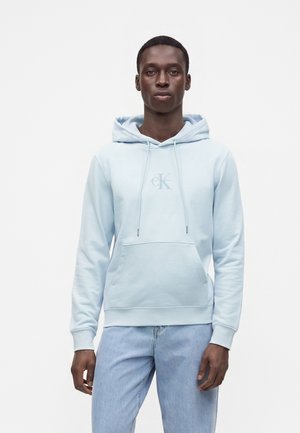 Young man wearing a light blue Calvin Klein hoodie and light blue jeans, standing against a plain white background.