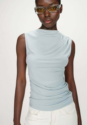 Jeune femme noire portant des lunettes rectangulaires à verres jaunes, un haut sans manches bleu clair et un pantalon blanc, sur un fond uni.