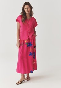 Blusa rosa a maniche corte con bottoni abbinata a una lunga gonna rosa con motivi floreali multicolori. Tessuto morbido e leggero.