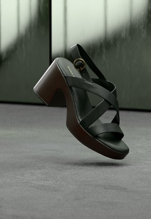 Μαύρο σανδάλι πλατφόρμα με σταυρωτούς ιμάντες και χρυσή πόρπη, εμφανίζεται να αιωρείται πάνω από γκρι μοκέτα με θολό φόντο παράθυρου.