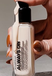 Bouteille de fond de teint en verre transparent avec une pompe noire ; le fond de teint liquide de couleur beige clair s'écoule du dessus ; étiquettes avec texte noir.