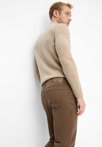 Beige strikket sweater med lange ærmer og tætsiddende manchetter. Mørkebrune bukser med syningsdetaljer og mærkelabel ved linningen. Ensfarvet hvid baggrund.