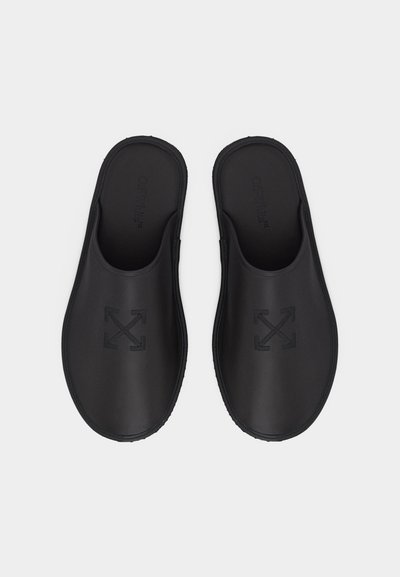 Ζευγάρι μαύρα λαστιχένια clogs χωρίς κορδόνια με ανάγλυφο λογότυπο διασταυρωμένων βελών στην κορυφή, φωτογραφημένα από πάνω σε λευκό φόντο.