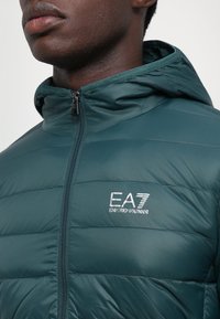 Grøn polstret jakke med hætte, der har en horisontal quiltet design, lynlåslukning og broderet EA7-logo på brystet. Glat tekstur.