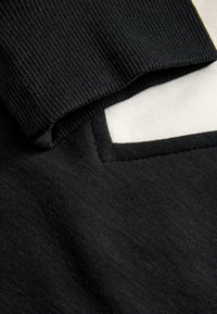 Schwarzer Strickstoff mit weißer Unterseite und kontrastierendem Besatz. Am Rand eine gerippte Textur und ein rechteckiges Taschen-Detail.