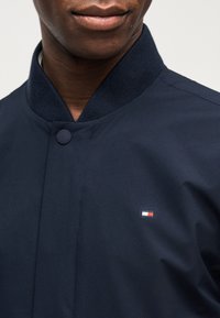 Κοντινό πλάνο ενός ατόμου που φοράει σκούρο μπλε σακάκι με ριμπ κοντό λαιμό και μικρό ορθογώνιο κόκκινο, λευκό και μπλε λογότυπο στο στήθος.