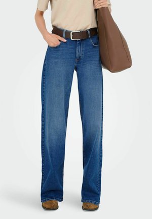 Blå vida jeans i denim med ljus tvätt, prydda med ett brunt bälte och en brun handväska, tillsammans med en beige skjorta.