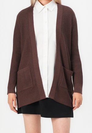 Žena v hnědém žebrovaném svetru s otevřeným předním dílem a kapsami, přes nějž má bílou košili na zapínání a černou sukni.