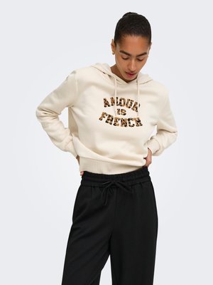 Person iført en cremefarvet hættetrøje med teksten "Amour is French" i leopardprint og sorte snor-bukser, som kigger ned med hænderne på hofterne.