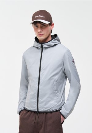 Ung mand iført en lysegrå hættejakke med en sort lynlås, brune bukser og en brun og hvid baseballkasket.