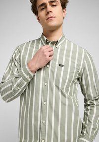 Camicia button-up verde con strisce verticali bianche, dotata di taschino sul petto, colletto button-down e bottoni in plastica.